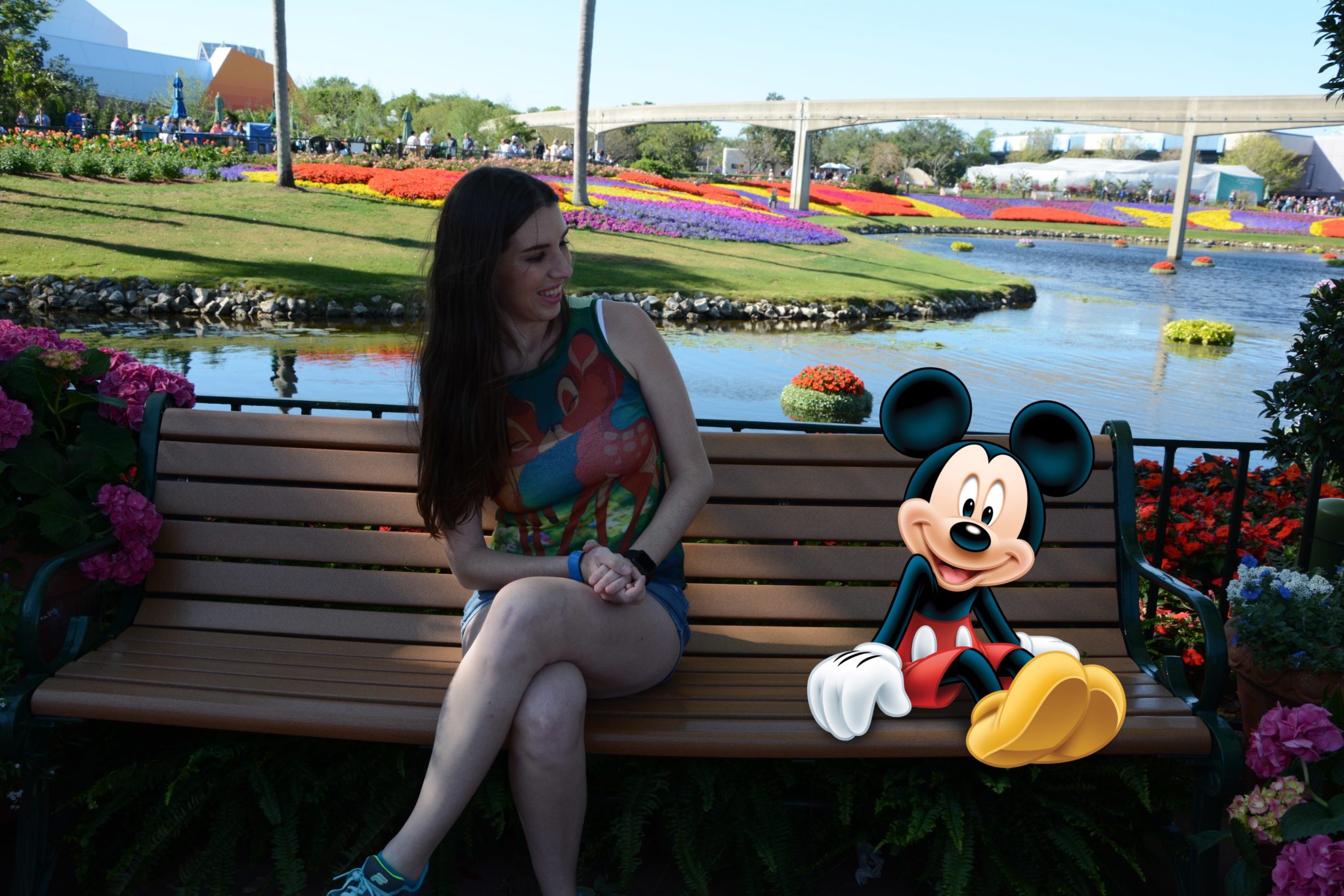 Chelsea sitting on a bench with Mickey Mouse at Epcot in front of the flower beds.
